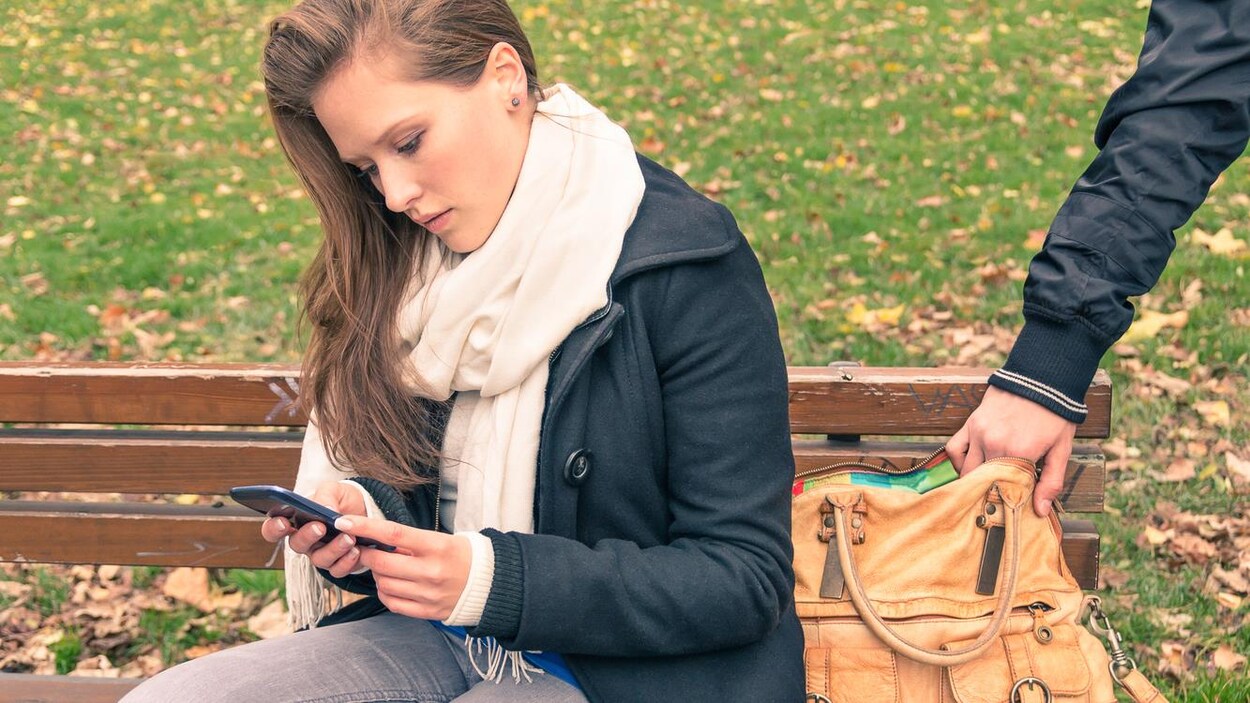 Une jeune femme assise sur un banc à l'extérieur regarde son téléphone portable. Pendant ce temps, on voit le main d'un voleur se glissant dans son sac à main. Une jeune femme assise sur un banc à l'extérieur regarde son téléphone portable. Pendant ce temps, on voit le main d'un voleur se glissant dans son sac à main.