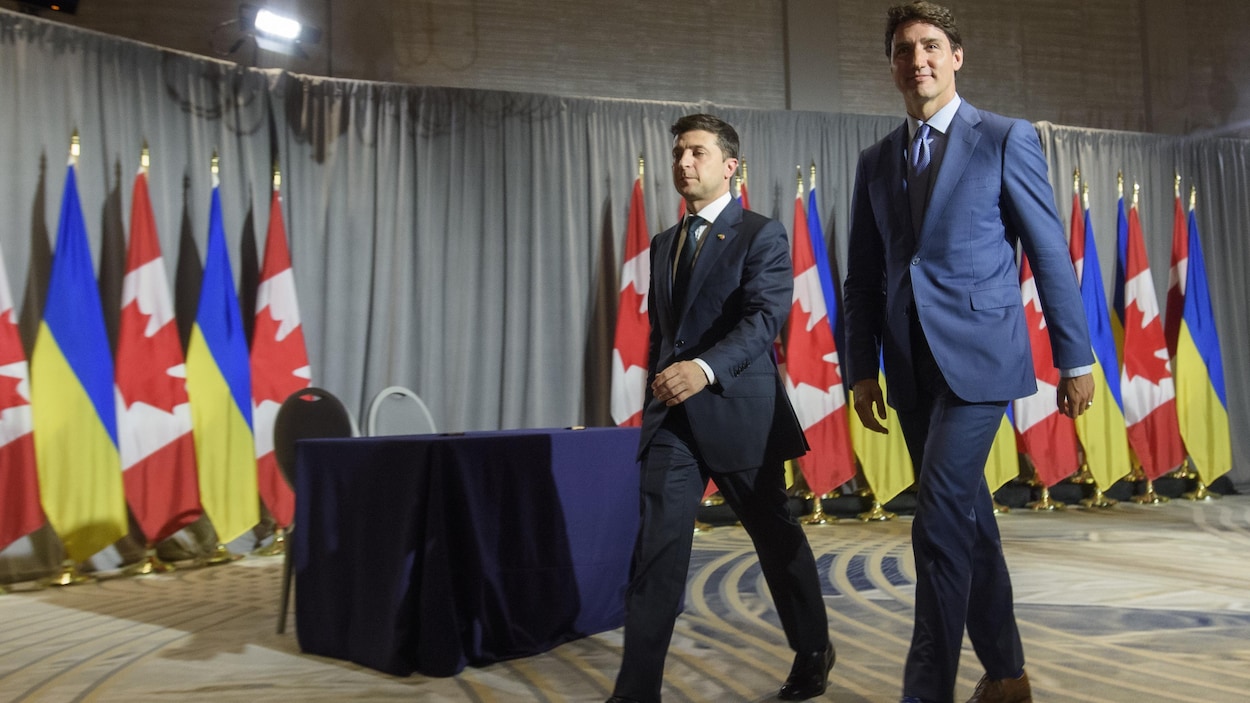 Les deux dirigeants marchent dans une pièce, derrière eux se trouvent des drapeaux de l'Ukraine et du Canada.