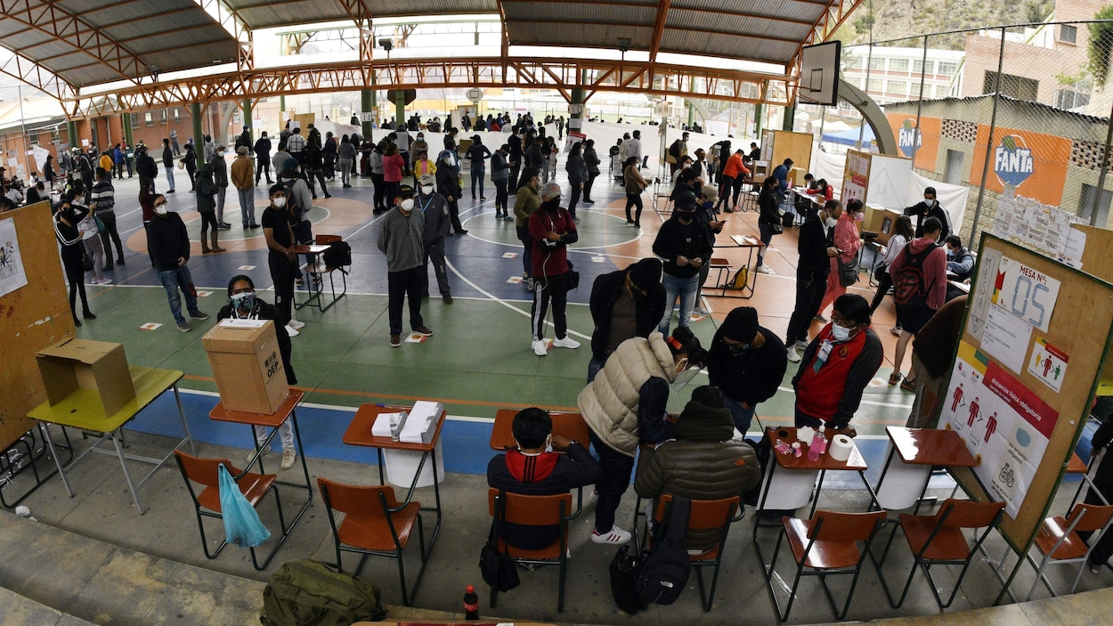 Des gens attendent en file, d'autres votent dans un gymnase extérieur couvert d'un toit.