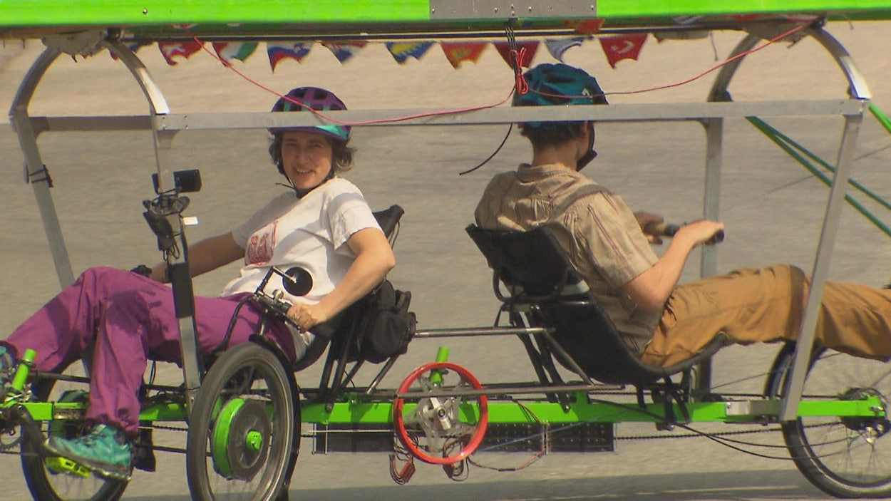 Une femme et un homme sont assis sur un vélo qui possède un écran solaire sur le toit. Elle est devant et doit pédaler, lui est derrière et doit ramer pour faire avancer le vélo.