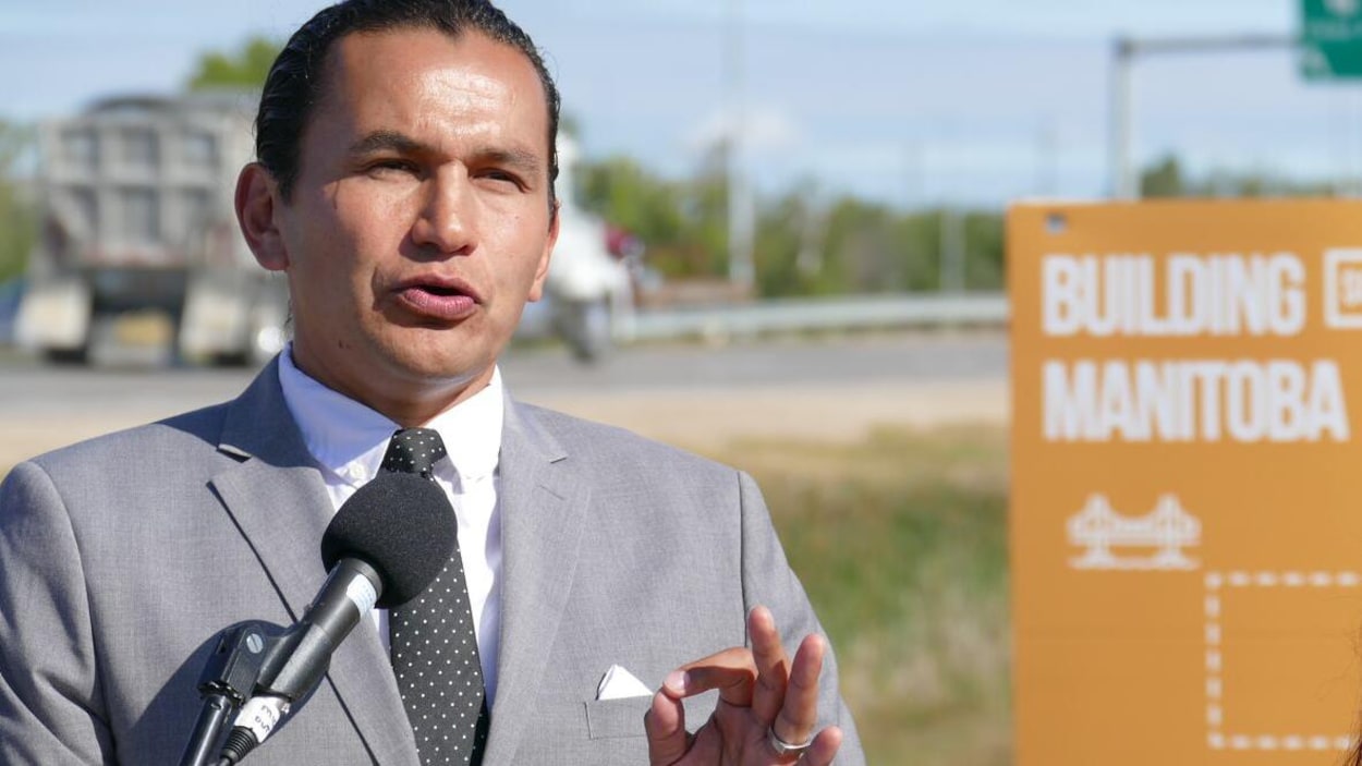 Un homme parle à un micro, à l'extérieur, devant une affiche orange aux couleurs du Nouveau Parti démocratique sur laquelle on peut lire en anglais : Construire le Manitoba.