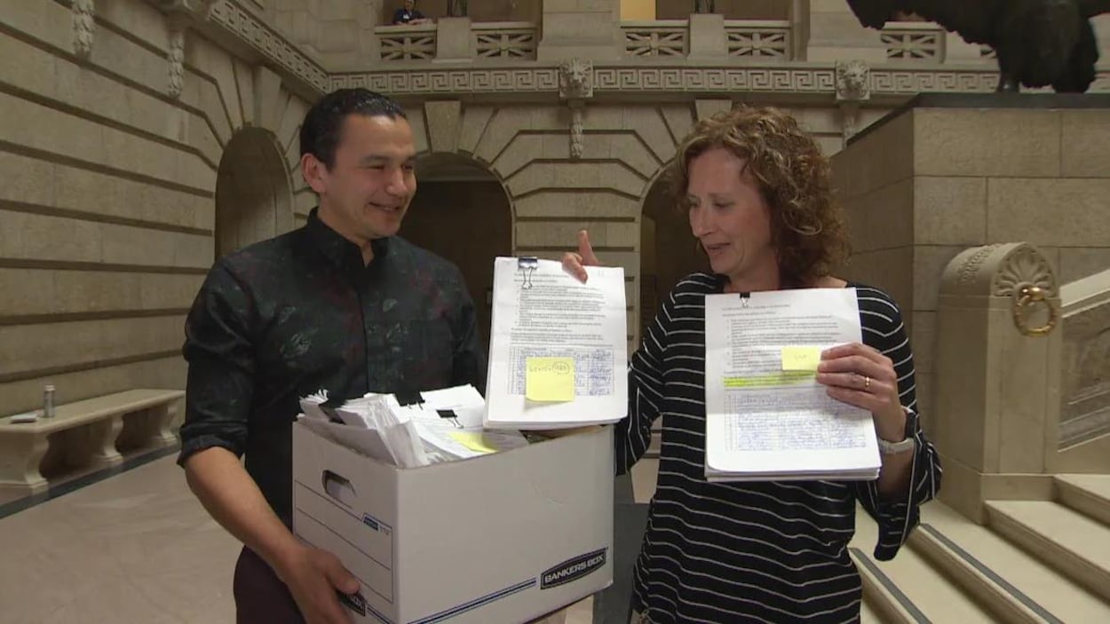 Un homme, Wab Kinew, tient une boite pleine de papiers, et une femme à ses côtés, Jodie Kehl, montre des pages de pétition portant des signatures. Ils sont tous deux dans le hall du Palais légslatif du Manitoba.