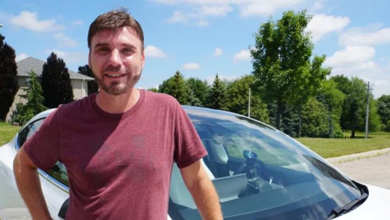 Wade Anderson à Woodstock, en Ontario, après 16 jours de voyage, devant sa voiture. Il a les cheveux châtains, une courte barbe, il porte un chandail bordeau