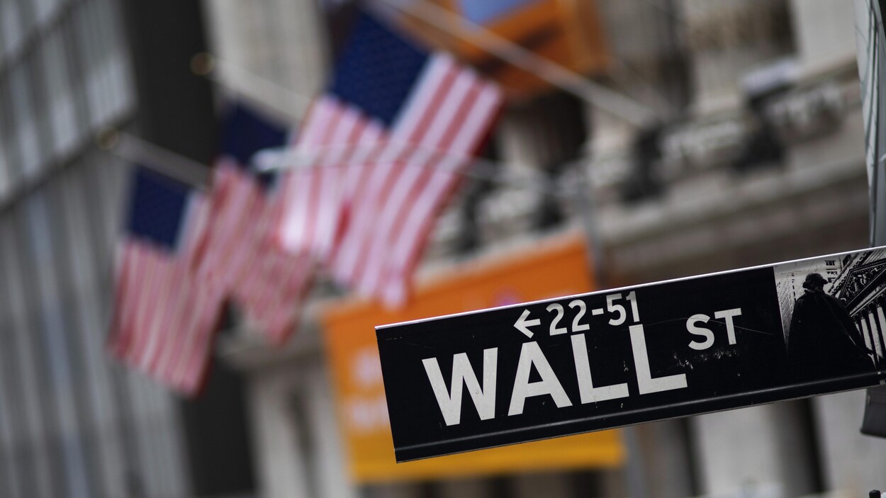 Le panneau de Wall Street photographié en gros plan avec, à l'arrière, une série de drapeaux américains suspendus à la façade de l'immeuble de la bourse, à New York.