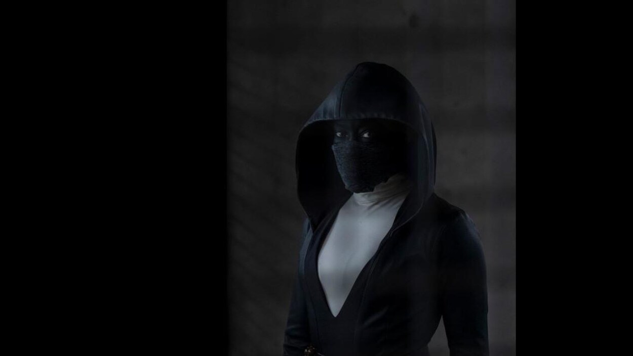 Une femme masquée porte un manteau noir avec un grand capuchon sur la tête. Elle regarde droit dans l'objectif de la caméra.