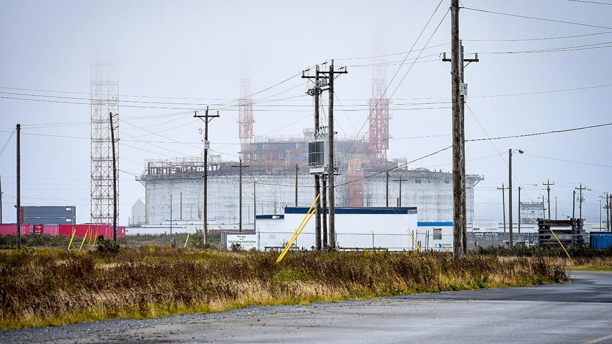 Un grand bâtiment en construction, entouré d'échafauds dont le sommet se perd dans la brume, vu depuis une route voisine.