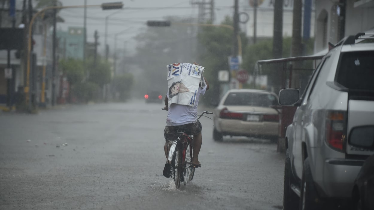 Un homme circule sur un vélo dans une rue recouverte par quelques centimètres d'eau. Le temps est lourd et il pleut.