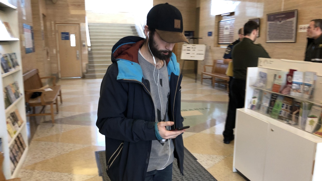 Un homme sort du palais de justice en regardant son téléphone, la tête baissée sous une casquette.