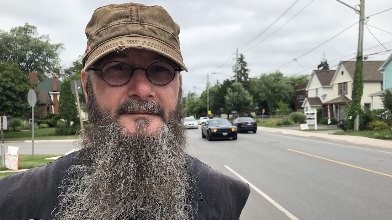 Un homme avec des lunettes rondes, une casquette sur la tête et une longue barbe regarde la caméra. On voit une rue qui s'allonge derrière lui.