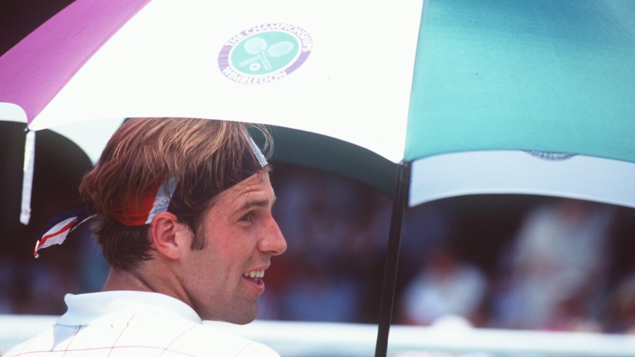 Un joueur de tennis, avec un bandeau multicolore dans les cheveux, se protège avec un parapluie aux couleurs du tournoi de Wimbledon.