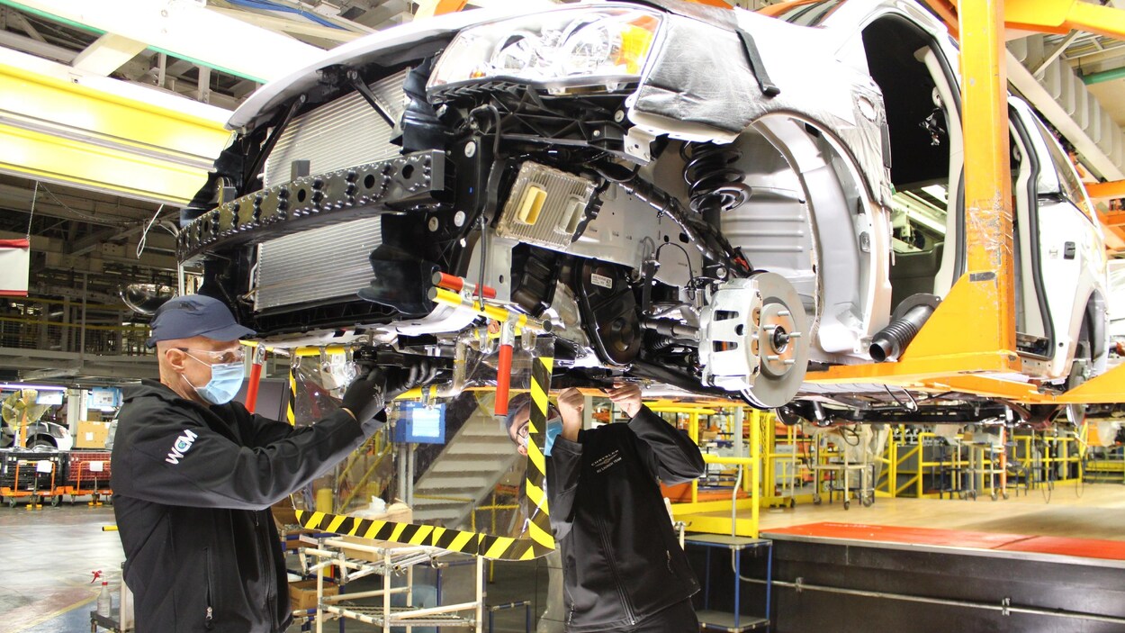 Deux ouvriers travaillent sous une fourgonnette Dodge Caravan dans une usine.