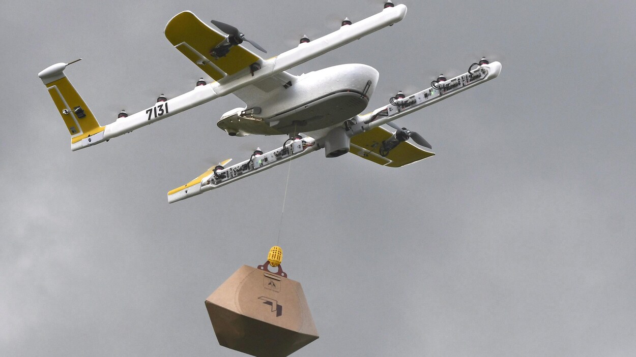 Une photo d'un drone ressemblant à un avion sous lequel un petit treuil retient un colis suspendu au bout d'un fil de fer.