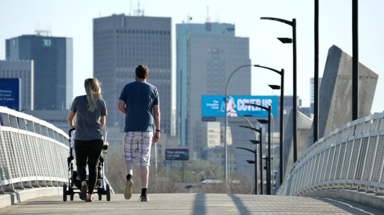 Un couple vu de dos promène une poussette sur un pont pour piétons, et les édifices du centre-ville de Winnipeg apparaissent en arrière-plan de la photo.