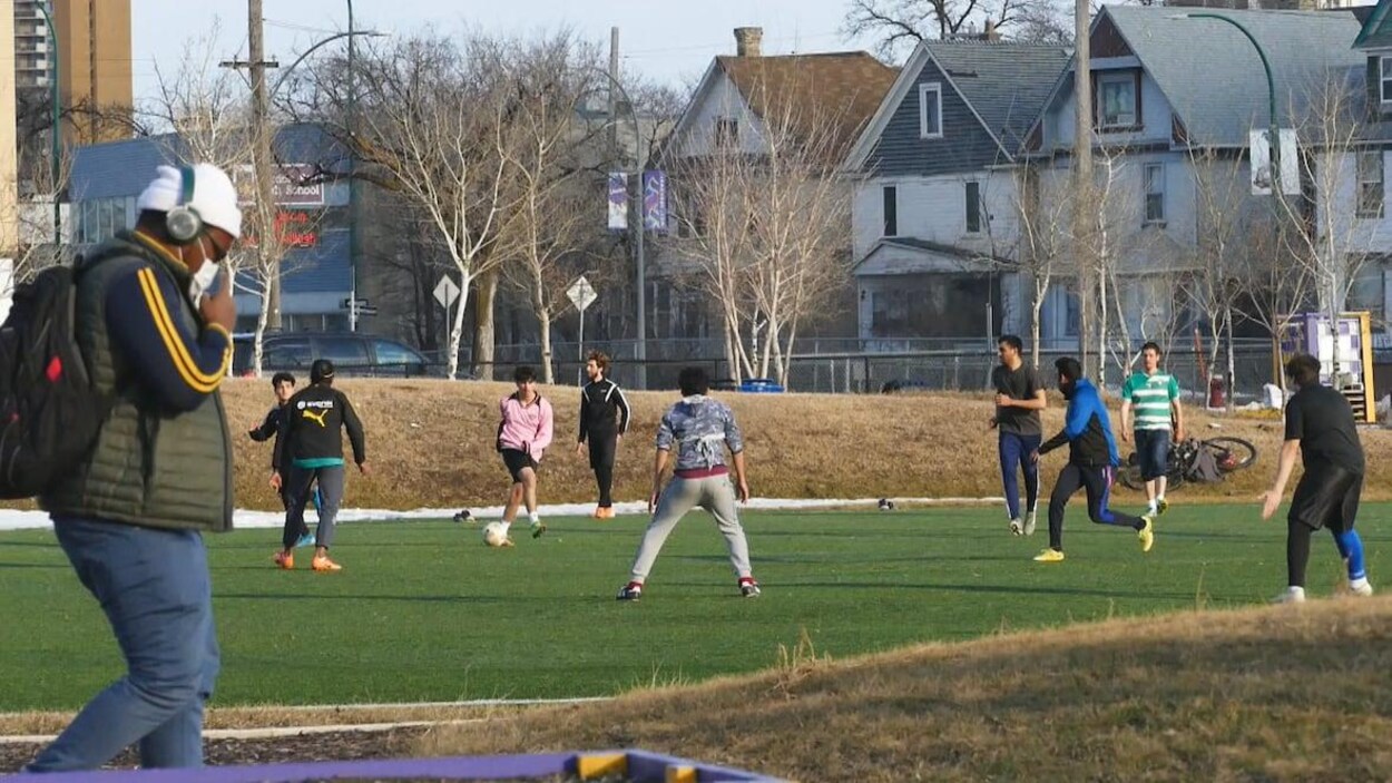 Une dizaine de jeunes jouent au soccer, alors qu'un homme portant un masque passe devant le terrain.