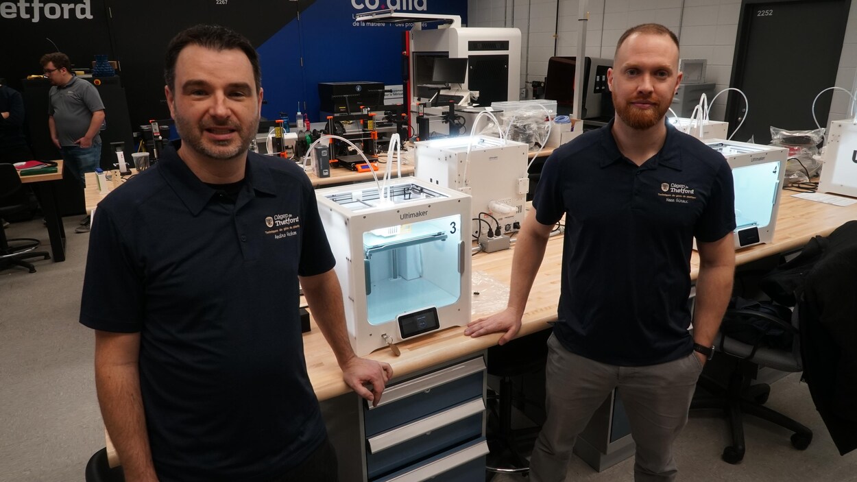 Deux hommes avec un chandail bleu se tiennent debout de chaque côté d'un imprimante 3D.
