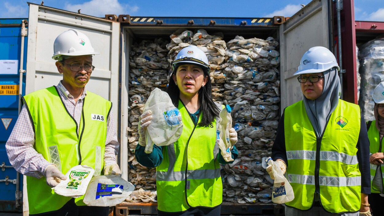 La ministre tient des bouteilles de plastique souillées dans ses mains, comme deux personnes qui se tiennent à côté d'elle. Ils sont revêtus d'habits de protection et des conteneurs remplis de déchets se trouvent derrière eux.