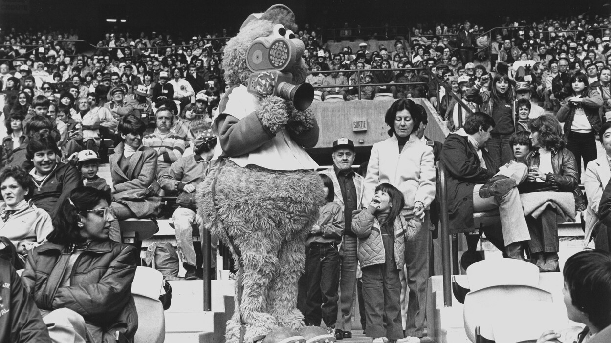 Mascotte Youppi entourée de la foule dans le stade tient une caméra de Radio-Canada.