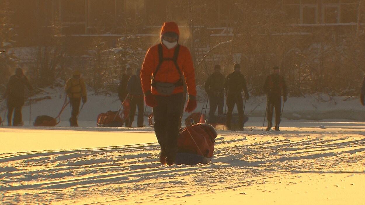Huit personnes marchent dans la neige en s'aidant de bâtons et en transportant de l'équipement dans leurs sacs à dos.