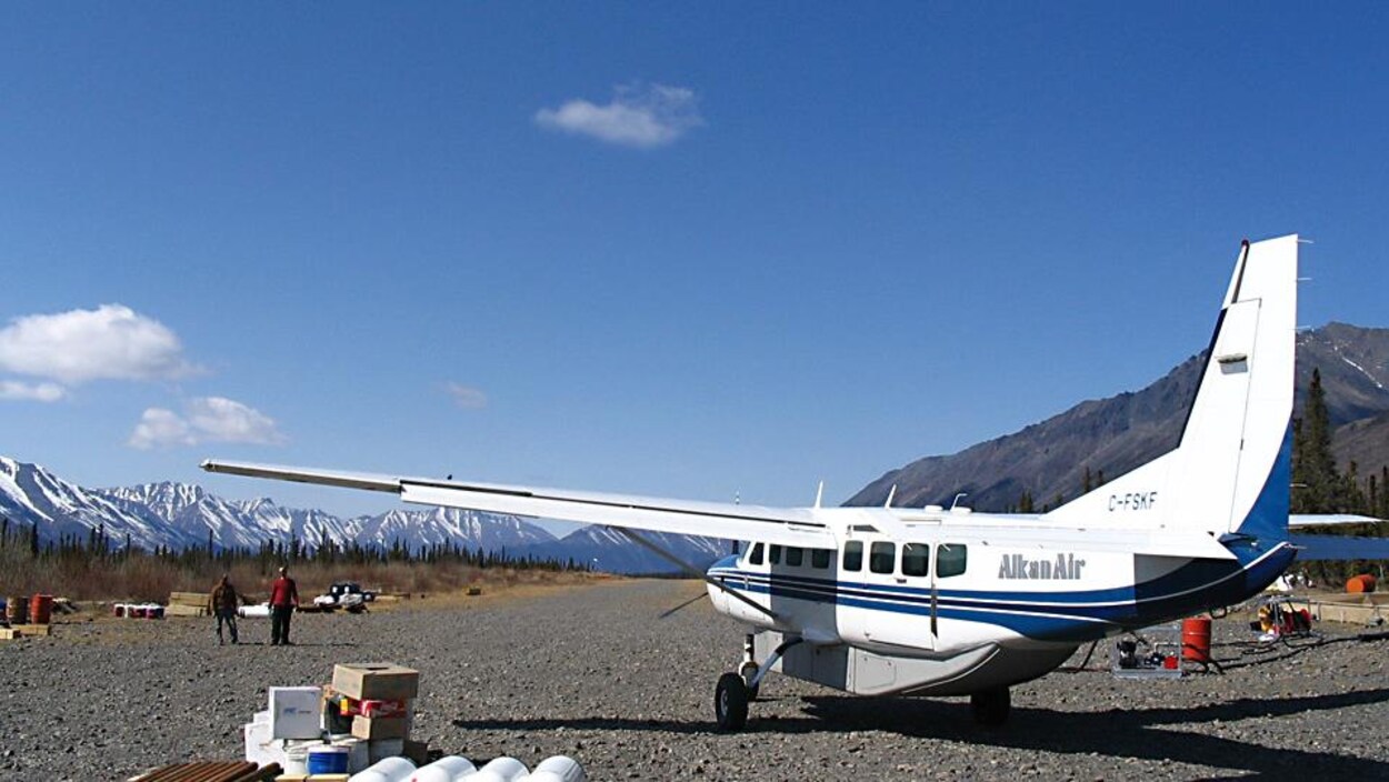 Un avion sur une piste d'atterrissage devant des montagnes et deux personnes qui marchent.