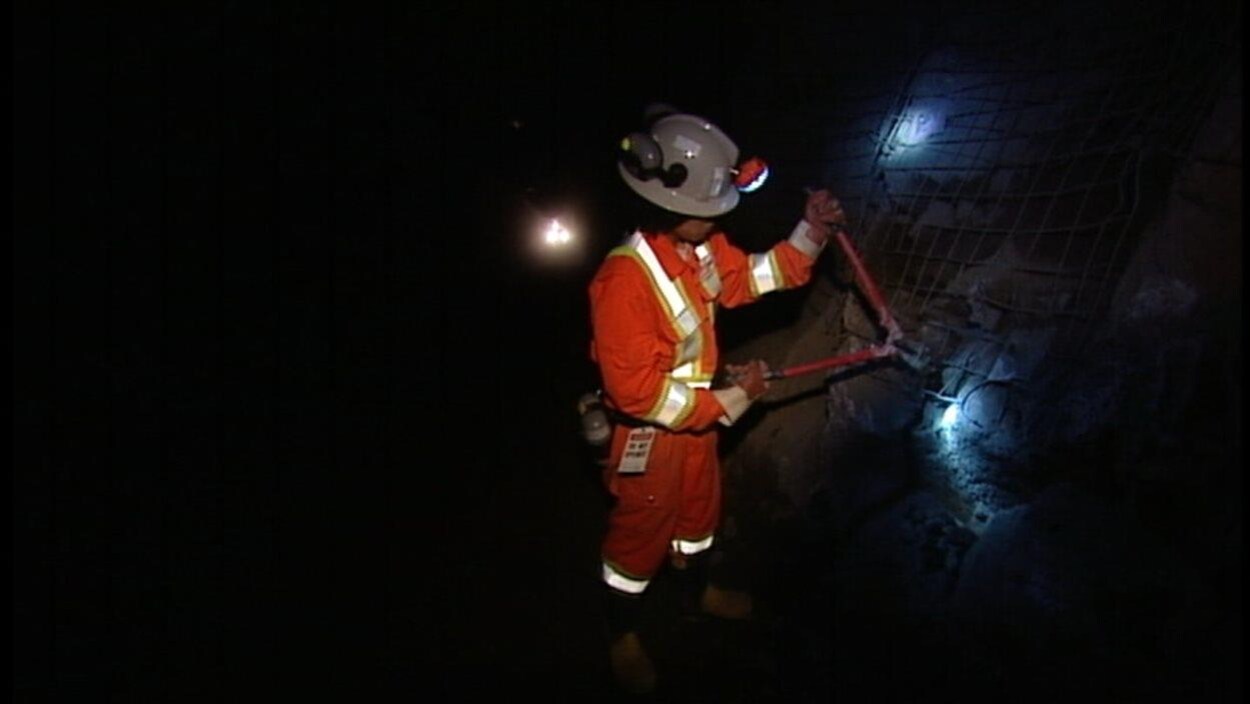 Un mineur en habit de sécurité orange installe un grillage à l'aide d'une lampe frontale.