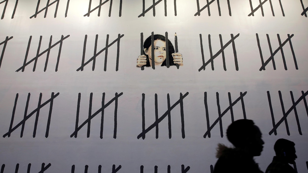 Deux passants marchent devant un mur blanc. Le visage de la journaliste et artiste Zehra Dogan y a été dessiné par Banksy derrière des barreaux, dont un est aussi un crayon. D'autres traits noirs représentent les jours passés en prison par la Turque. Deux passants marchent devant un mur blanc. Le visage de la journaliste et artiste Zehra Dogan y a été dessiné par Banksy derrière des barreaux, dont un est aussi un crayon. D'autres traits noirs représentent les jours passés en prison par la Turque.