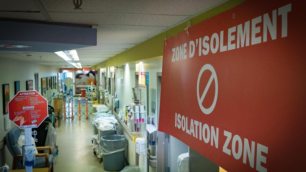 On voit en plongée un couloir d'hôpital. En gros plan, à droite de l'image, on voit un grand écriteau en rouge où il est écrit « zone d'isolement » avec le symbole d'interdiction de passer. On voit en plongée un couloir d'hôpital. En gros plan, à droite de l'image, on voit un grand écriteau en rouge où il est écrit « zone d'isolement » avec le symbole d'interdiction de passer.