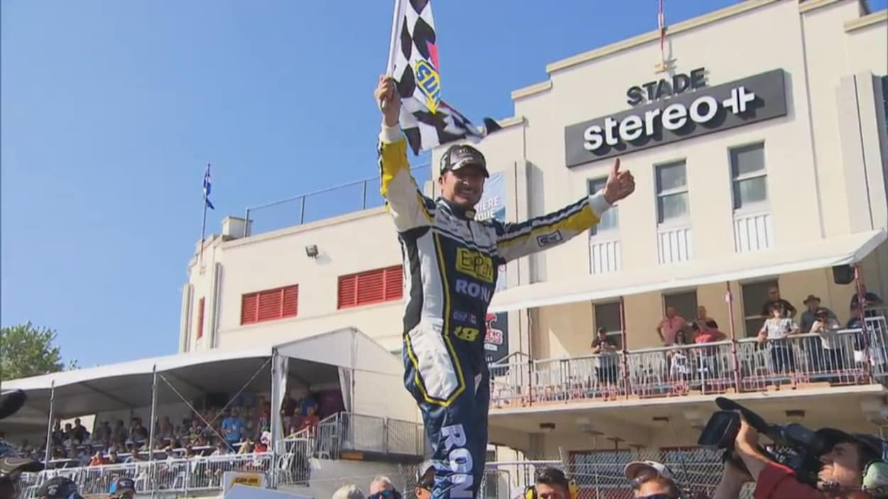 Un pilote de course automobile sourit et tient le drapeau à damiers d'une main sur le toit de son véhicule. Un pilote de course automobile sourit et tient le drapeau à damiers d'une main sur le toit de son véhicule.
