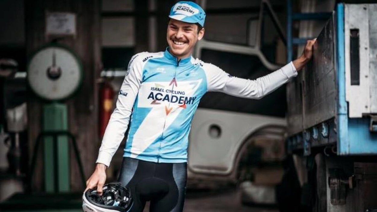 Il est appuyé sur un camion et porte maillot et casquette aux couleurs d'Israel Cycling Academy.
