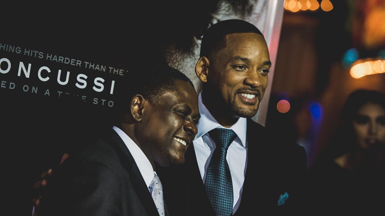 Dr Bennett Omalu et Will Smith se tiennent côte à côte et sourient aux caméras devant une affiche du film Commotion. Dr Bennett Omalu et Will Smith se tiennent côte à côte et sourient aux caméras devant une affiche du film Commotion.
