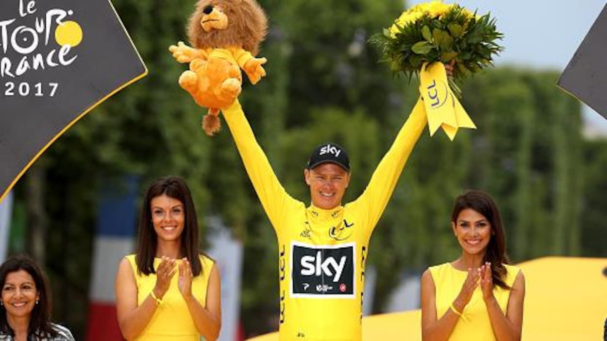 Chris Froome, tout sourire, accompagné de deux hôtesses sur le podium des Champs-Élysées en 2017. Chris Froome, tout sourire, accompagné de deux hôtesses sur le podium des Champs-Élysées en 2017.