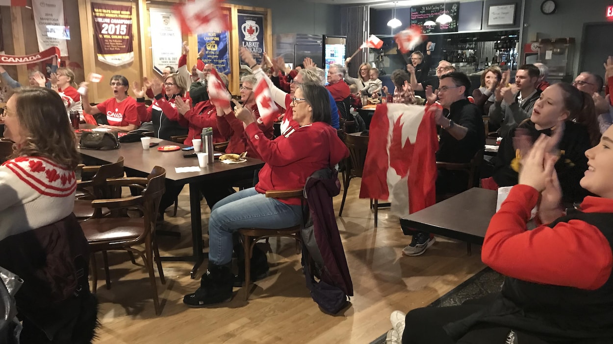 Une foule de gens assis à des tables sont habillés en rouge, brandissent des drapeaux canadiens et applaudissent. Une foule de gens assis à des tables sont habillés en rouge, brandissent des drapeaux canadiens et applaudissent.