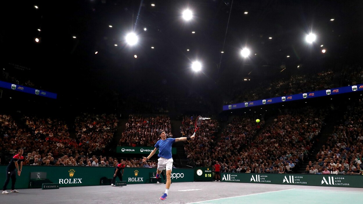 Un joueur de tennis retourne une balle en sautant. Il est devant des gradins bondées.