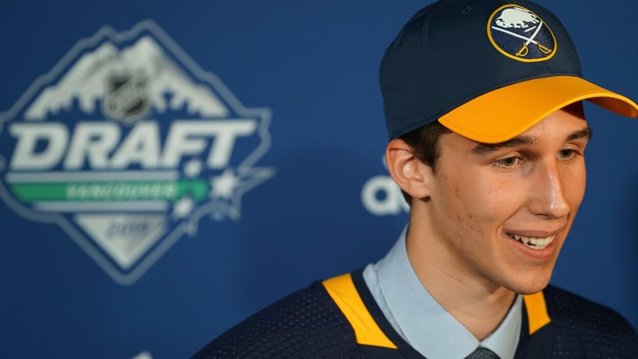 Un jeune homme souriant pose de profil avec une casquette de sport et un chandail d'hockey.