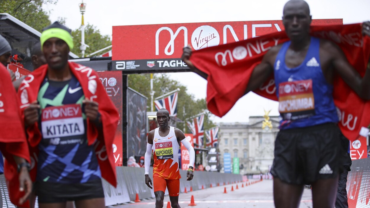 Un coureur vêtu de blanc et rouge franchit le fil d'arrivée pendant que le vainqueur célèbre sa victoire.