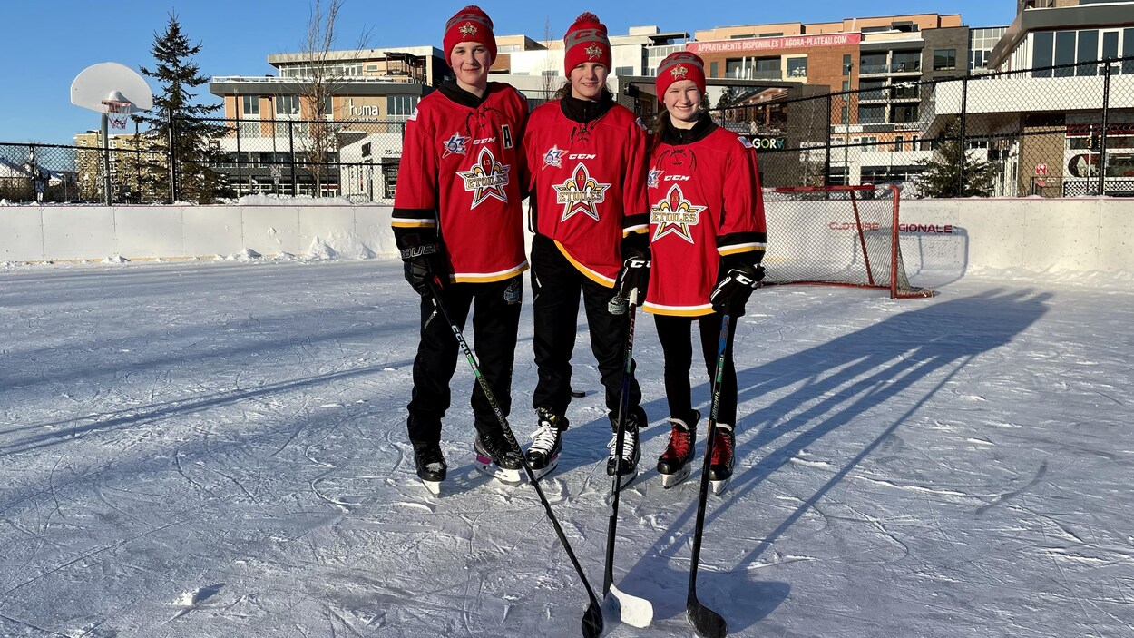 Trois jeunes femmes prennent une photo avec leur bâton de hockey sur une patinoire extérieure.