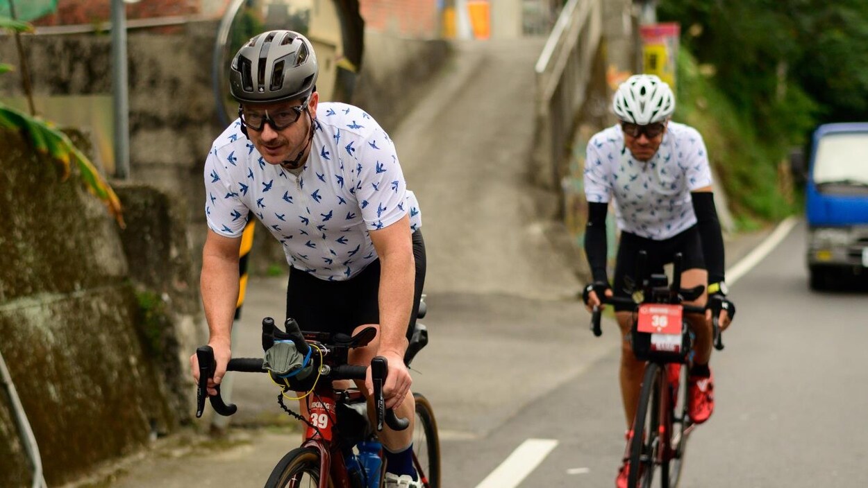 Deux hommes sont à vélo dans une côte de Taïwan. Ils portent le même ensemble, blanc avec des oiseaux bleus.