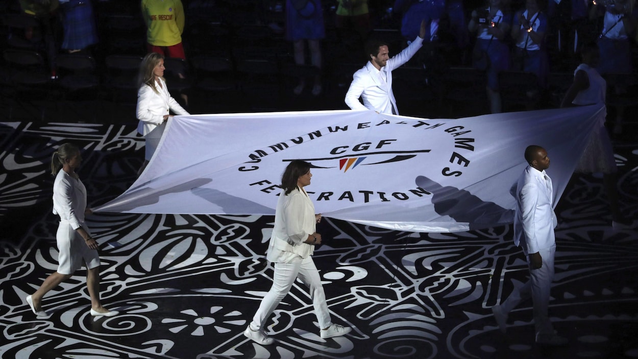 Six personnes défilent avec un grand drapeau blanc de la Fédération des Jeux du Commonwealth.
