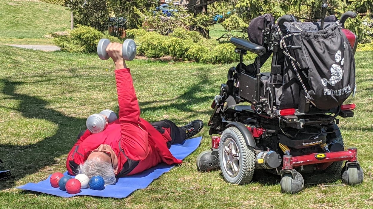 Il est couché au sol avec des haltères, à côté de son fauteuil motorisé.