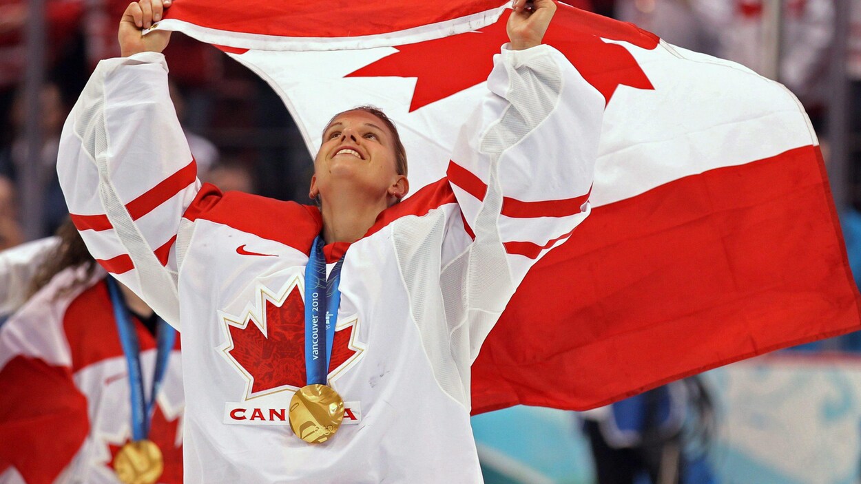 Une gardienne de but au hockey fait flotter le drapeau canadien au-dessus de sa tête. Elle a une médaille d'or autour du cou.