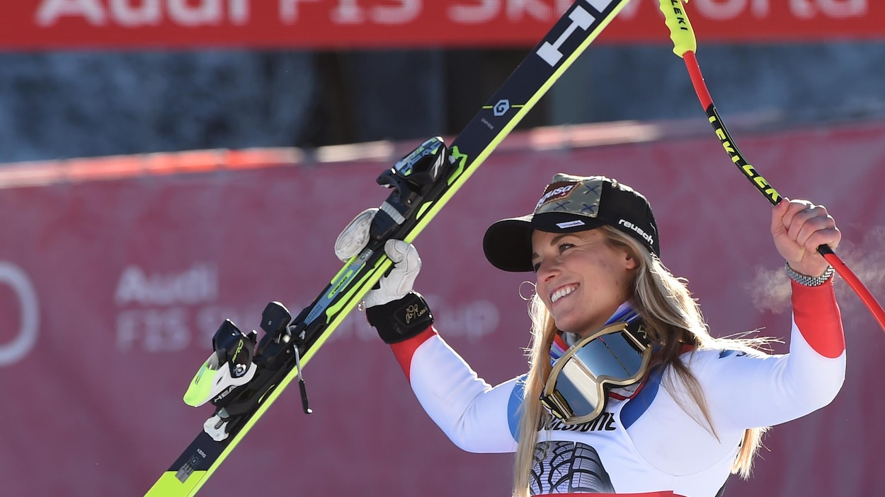 Elle sourit et tient un bâton dans sa main gauche et un ski dans celle de droite.