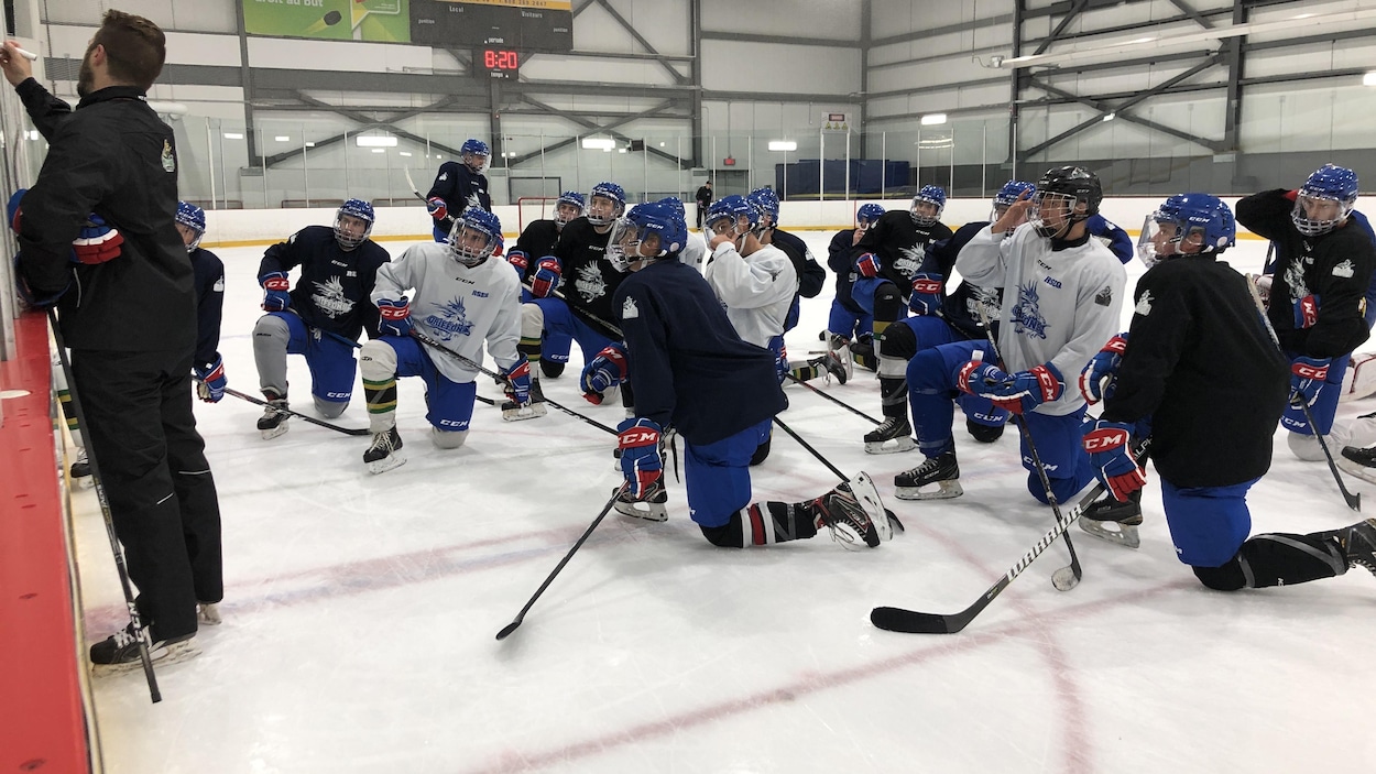Un entraîneur montre des exercices sur un tableau à des joueurs agenouillés sur la patinoire.