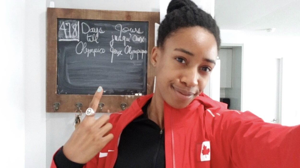 Une femme sourit et pointe par-dessus son épaule un tableau indiquant le nombre de jours d'ici les Jeux olympiques de Tokyo : 478.
