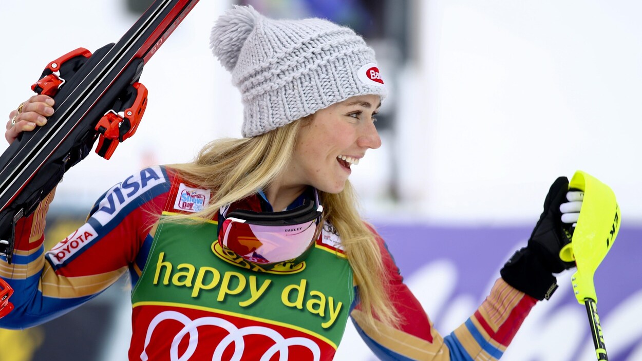 Elle sourit avec ses skis dans la main droite.