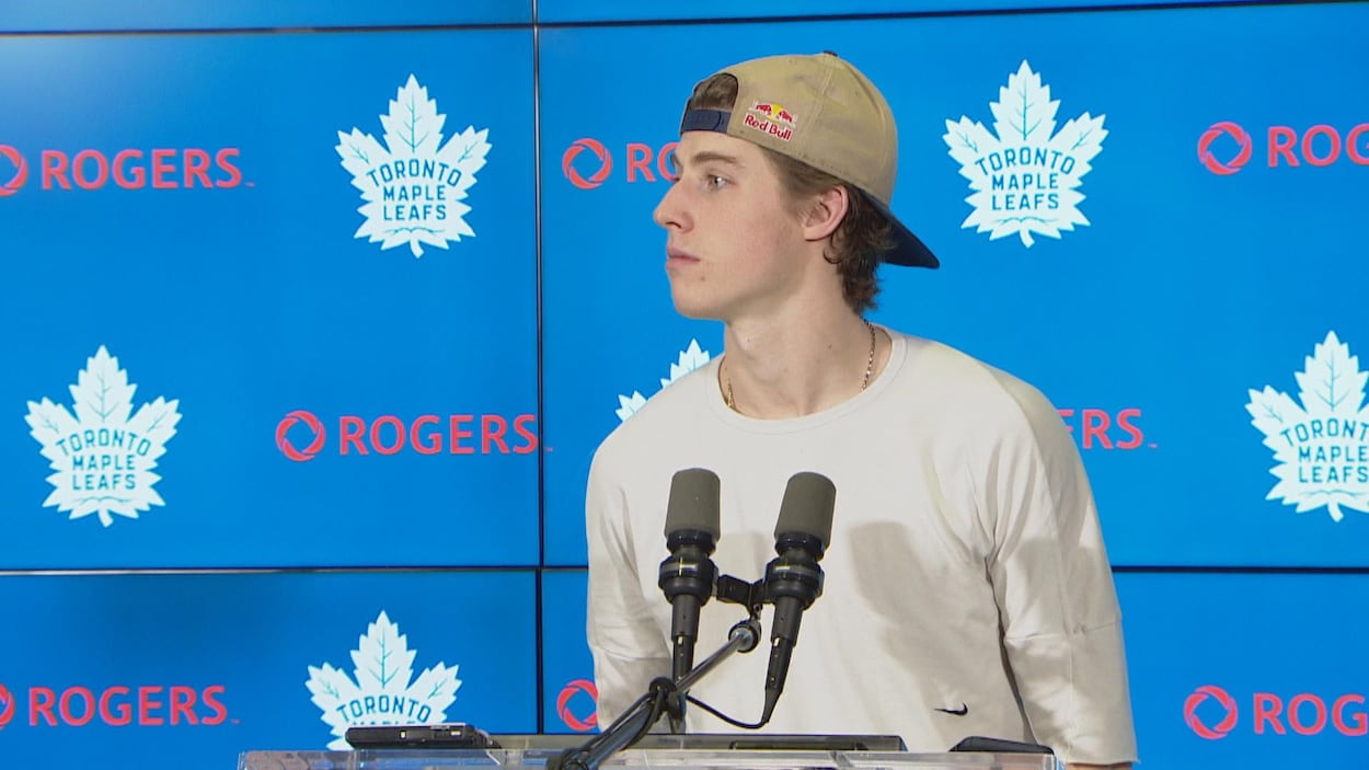 Un homme porte une casquette. Il écoute attentivement une question d'un journaliste.