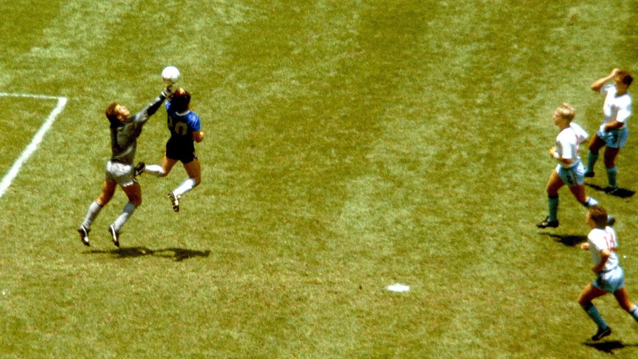 Un joueur en bleu saute devant un gardien et frappe le ballon de sa main gauche