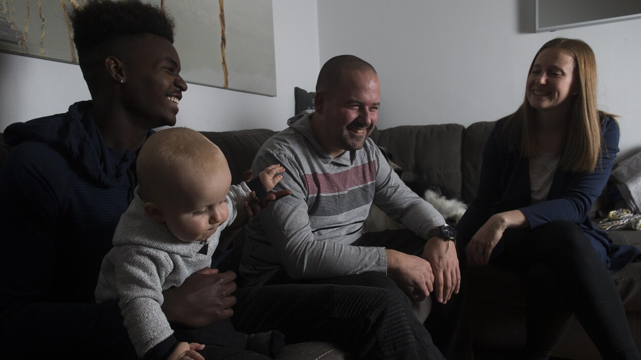 Prinzy Dessources, qui tient dans ses bras le petit Madden, est assis sur un divan près de son père et de sa mère. Prinzy Dessources, qui tient dans ses bras le petit Madden, est assis sur un divan près de son père et de sa mère.