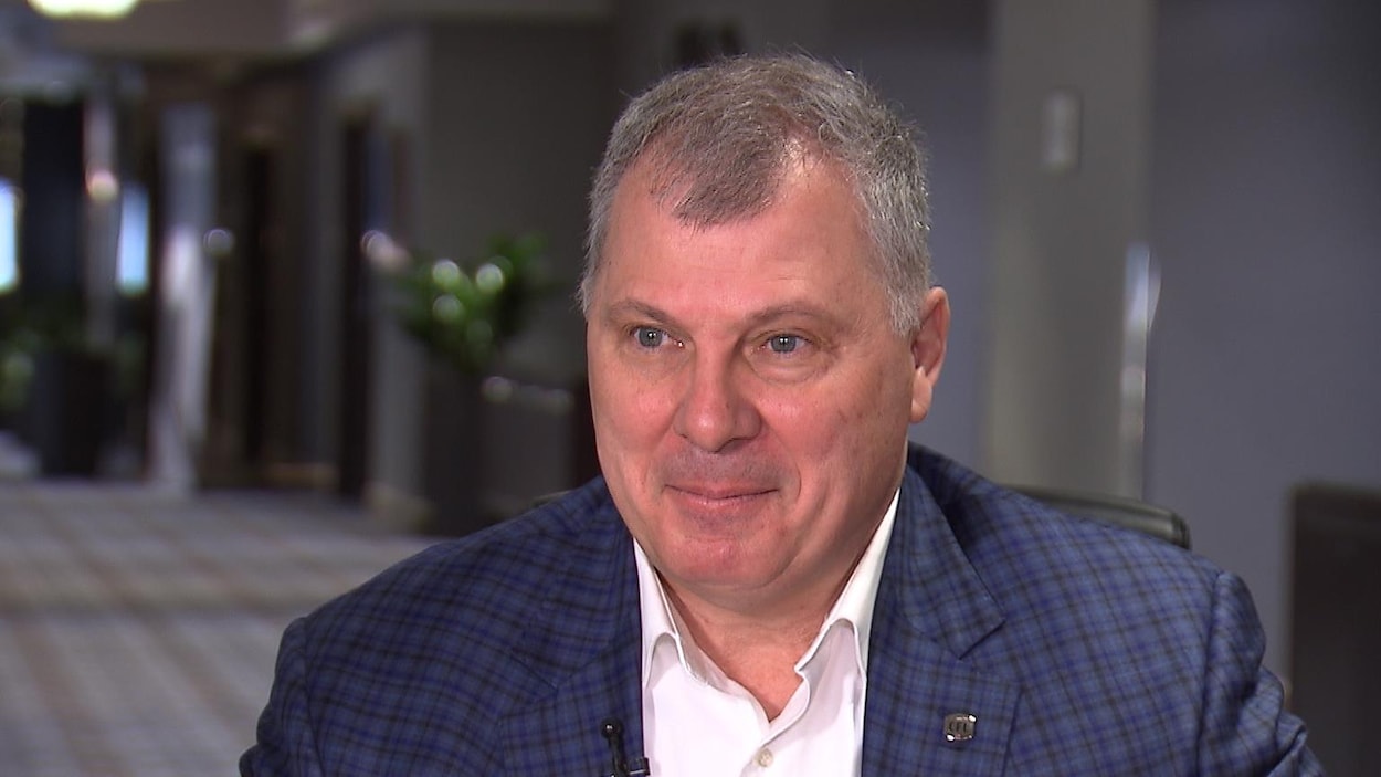 Un homme vêtu d'un costume mauve et noir et d'une chemise blanche est interviewé par un journaliste de Radio-Canada.