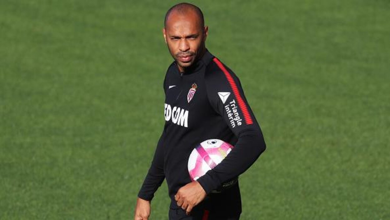 Il tient un ballon sous le bras le gauche sur un terrain d'entraînement.