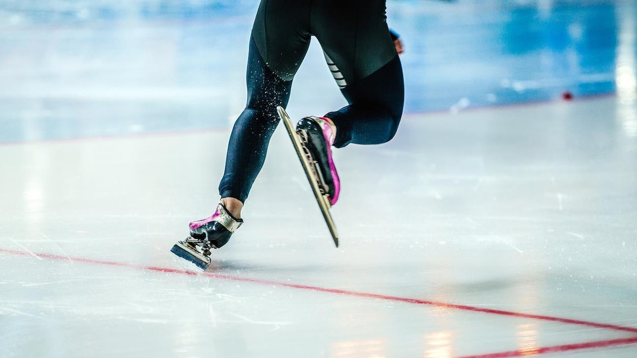 Un patineur de vitesse en action sur la glace lors du départ. On ne voit que ses patins et ses jambes. L'athlète porte des collants noirs.
