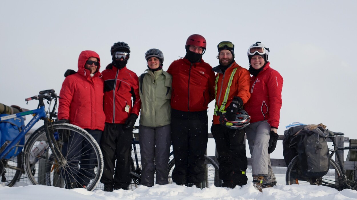 Nous voyons 6 cyclistes, l'hiver, souriants. Ils portent des manteaux d'hier et certains d'entre eux ont des casques et des lunettes de ski. Nous voyons quelques vélos à gauche et à droite ainsi que de la neige au sol. Le ciel est gris.